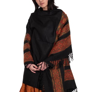 May include: A black shawl with a colorful geometric pattern and fringe trim. The shawl is draped over a person wearing a long orange skirt.