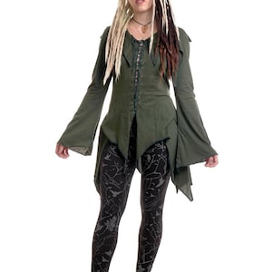 Può includere: Una tunica a maniche lunghe verde oliva con allacciatura frontale, orlo a punta e maniche svasate, abbinata a leggings neri con motivo bianco di uccelli e rami, e stivali con plateau scuri e fantasia con spessi lacci neri.