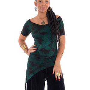 Könnte beinhalten: Ein grünes und schwarzes schulterfreies Oberteil mit Batikmuster, kurzen Ärmeln und asymmetrischem Saum. Das Oberteil wird mit einer schwarzen Hose mit weitem Bein kombiniert. Zu den Accessoires gehören Goldschmuck und ein Ring.
