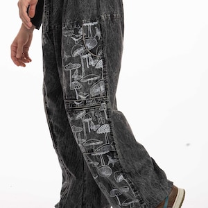 May include: Loose-fitting charcoal grey acid-wash trousers featuring a white line-art print of various mushrooms and butterflies. The trousers have a wide-leg silhouette with a side cargo pocket panel that displays the detailed botanical pattern.