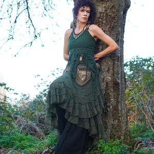 May include: A woman in a dark green layered skirt and top. The top has a halter neck and fringe. The skirt has multiple ruffles. She is leaning against a tree outdoors. The outfit is a bohemian style.