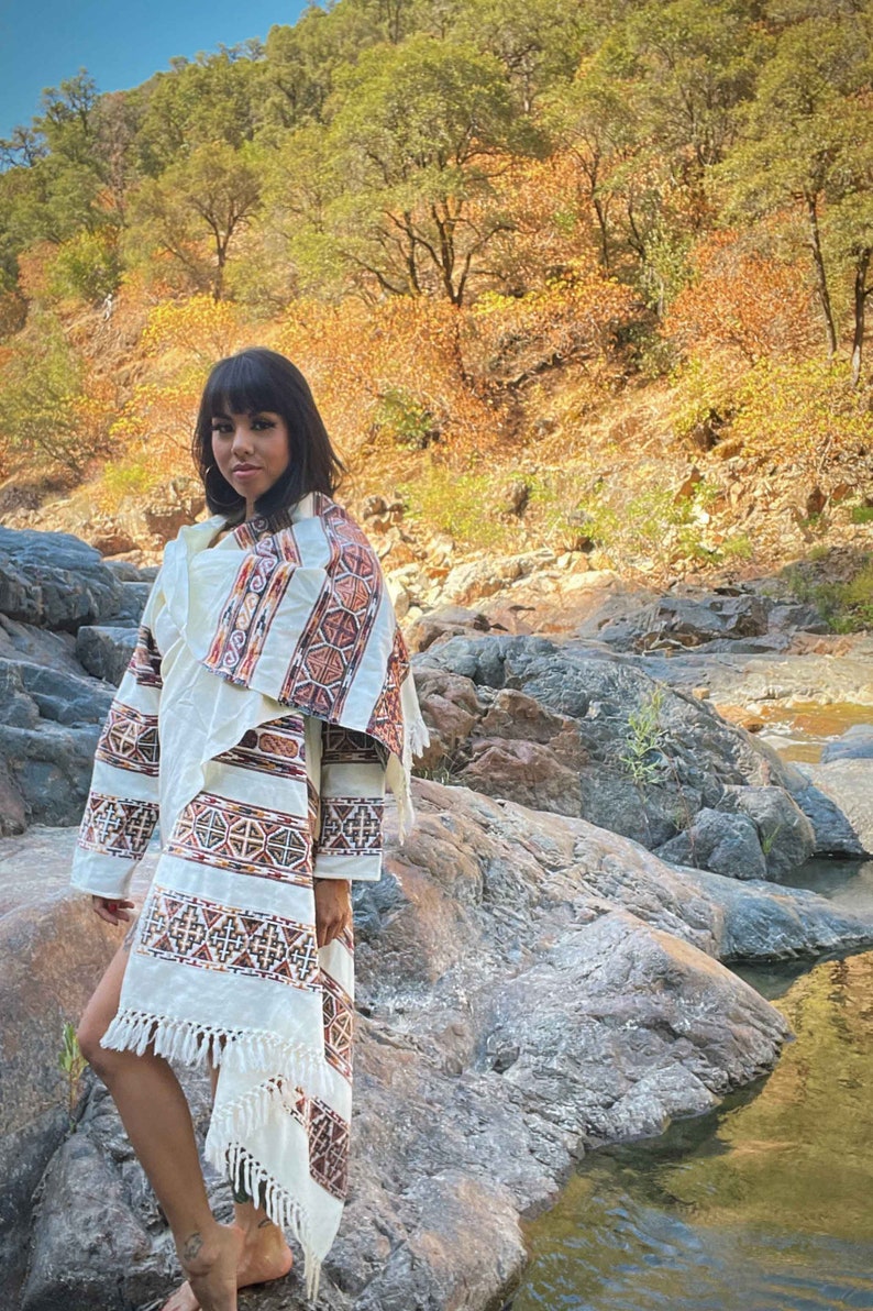 以下が含まれることがあります： フリンジ付きの白と茶色の模様のショールを着た人。ショールは人の肩にかけられ、腰で結ばれています。人は、背景に小川のある岩場の中に立っています。