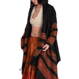 May include: A woman wearing a black shawl with red and gold trim over a white crop top and a long orange skirt with a floral pattern.