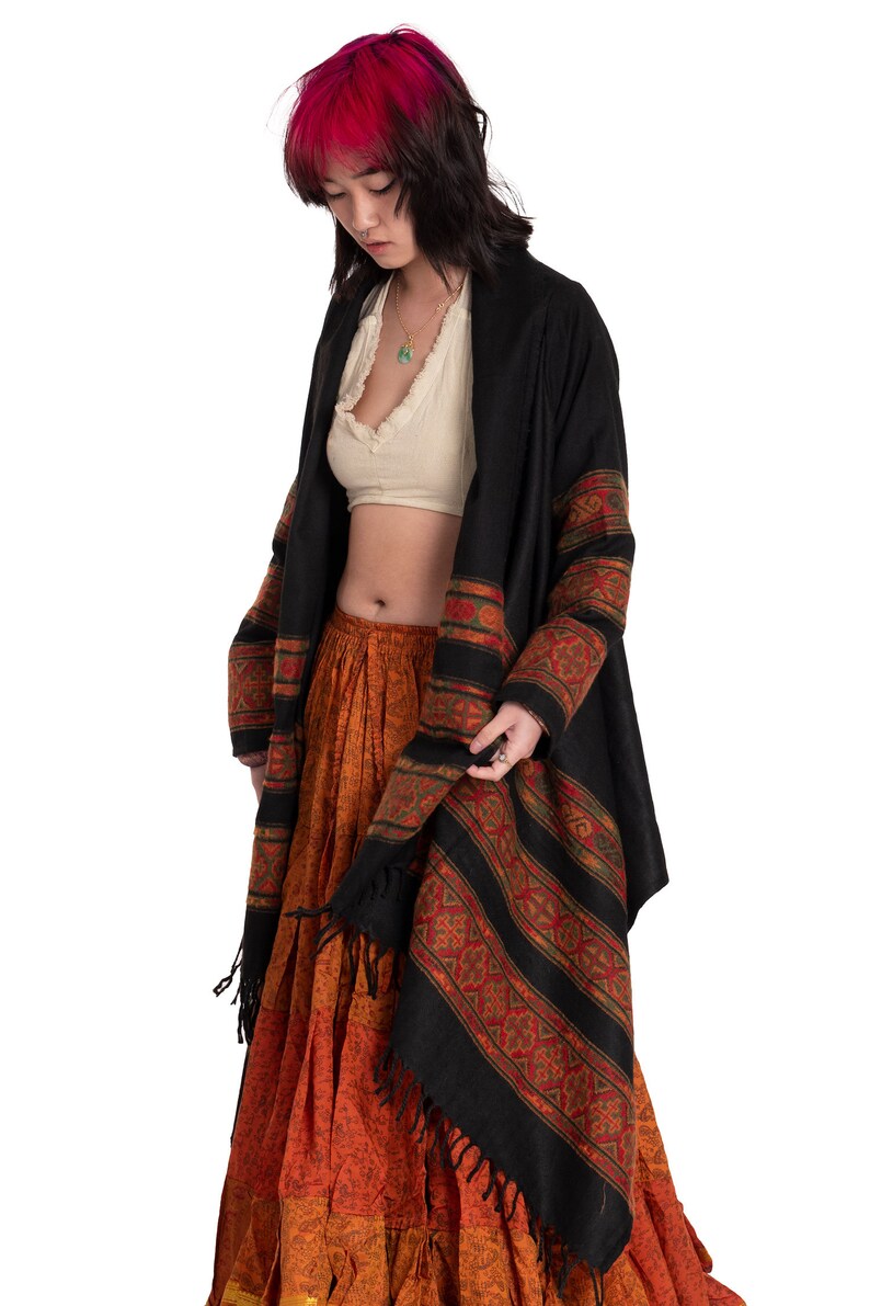 May include: A woman wearing a black shawl with red and gold trim over a white crop top and a long orange skirt with a floral pattern.