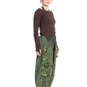 Könnte beinhalten: Ein braunes Langarm-Oberteil mit asymmetrischem Saum, kombiniert mit grünen Hosen mit Pilzaufdruck. Die Hose hat Taschen und einen Kordelzug in der Taille. Schwarze Stiefel vervollständigen das Outfit.