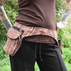 May include: A brown and beige patterned fabric belt with two attached pouches. The belt is worn over a brown tank top and black trousers.