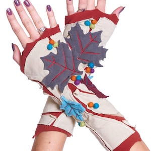 Può includere: Un paio di guanti senza dita bianchi con bordini rossi e decorazioni a forma di foglie in feltro. Le foglie sono grigie con cuciture rosse e hanno palline di feltro colorate attaccate. I guanti sono decorati con foglie e palline di feltro aggiuntive in vari colori.