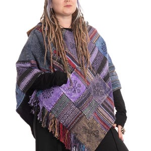 Könnte beinhalten: Ein Patchwork-Poncho in Blau-, Lila- und Brauntönen mit Fransen am Saum. Der Poncho weist verschiedene geometrische und florale Muster auf. Die Person trägt schwarze Langarmshirts und Hosen.