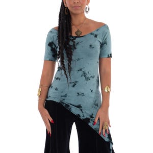 Puede incluir: Una blusa turquesa con teñido anudado, mangas cortas y dobladillo asimétrico. La blusa se combina con pantalones negros de pernera ancha. La modelo lleva joyas doradas, incluyendo un collar, pulseras y anillos. El fondo es blanco.