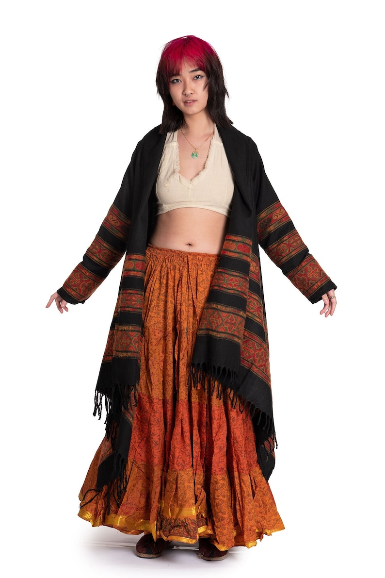 May include: A woman wearing a black and red patterned shawl over a white crop top and a long orange and brown skirt.