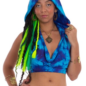 Könnte beinhalten: Ein blaues Kapuzen-Crop-Top mit Batikmuster und tiefem V-Ausschnitt. Das Oberteil wird mit einer schwarzen Hose kombiniert. Zu den Accessoires gehören goldener Schmuck wie Ohrringe, Halsketten, Armbänder und ein Ring. Das Model hat lange schwarze und grüne Zöpfe.