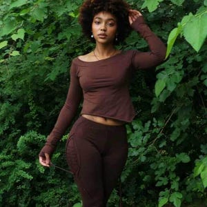 Può includere: Una maglia a maniche lunghe marrone scuro con scollo ampio e leggings marrone scuro abbinati con arricciature laterali e un pannello testurizzato simile al pizzo sull'orlo inferiore. Il completo è realizzato in tessuto a tinta unita.