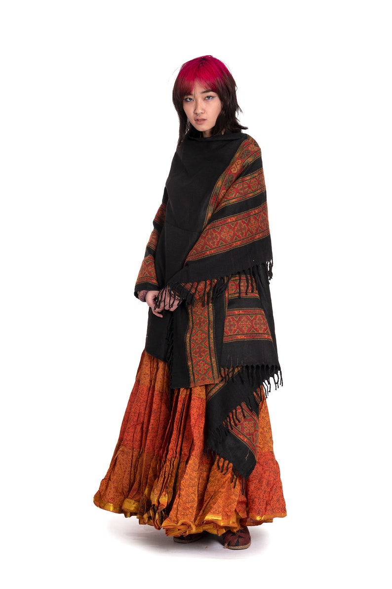 May include: A woman with pink hair wearing a black shawl with a colorful patterned border and a long orange and brown skirt.