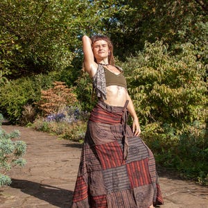 May include: A long, floor-length wrap skirt featuring a patchwork design with alternating panels of dark brown, black, and rust-red stripes, accented by circular floral patterns. The skirt includes a fabric tie at the waist for a secure fit.