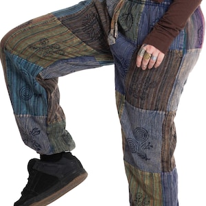 Op de afbeelding: Patchworkbroek in tinten blauw, groen en bruin met een tailleband met trekkoord. De broek heeft een verscheidenheid aan stoffen patches met patronen en wordt gedragen met zwarte sneakers. De algehele stijl is casual en bohemien.