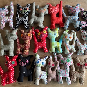 Könnte beinhalten: Eine Sammlung handgefertigter Stoffkatzenpuppen in verschiedenen Farben und Mustern. Jede Katze hat Knopfaugen und einzigartige Stoffdesigns, darunter Blumen-, Tupfen- und gemusterte Textilien. Die Katzen sind auf einer Holzoberfläche angeordnet.