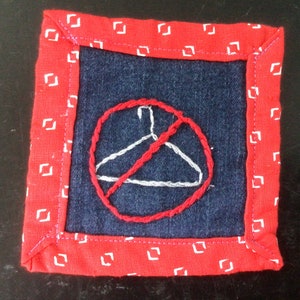 Peut inclure: Un carré de tissu rouge à pois blancs. Au centre du carré se trouve un carré de tissu denim bleu avec un cercle rouge et un cintre blanc barré d'une ligne rouge. Le texte "No Hangers" est brodé en blanc sur le denim.