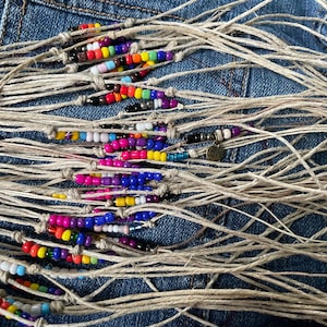 Könnte beinhalten: Nahaufnahme eines Hintergrunds aus Jeansstoff mit einer Sammlung bunter Perlenarmbänder, die mit natürlichem Garn zusammengebunden sind. Die Armbänder zeigen eine Vielzahl von Farben, darunter Blau, Lila, Pink, Gelb und Orange. Eines der Armbänder hat einen kleinen Anhänger mit der Aufschrift "Love".