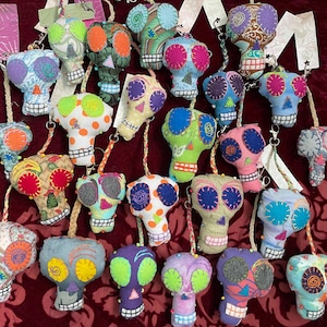 May include: A collection of colorful, handmade felt skull keychains. Each skull is decorated with different patterns and colors, including polka dots, stripes, and floral designs. The keychains are strung on colorful cords.