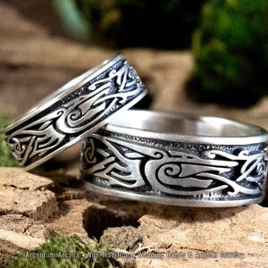 May include: Two silver wedding bands with a Celtic knot design featuring stylized birds. The bands are on a wooden surface with green moss.