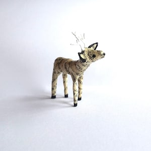 May include: A small, paper mache deer sculpture with white antlers and black eyes. The deer is covered in printed text from a book.