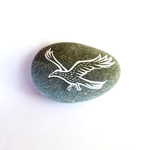 May include: A smooth, gray stone with a white outline of a flying bird. The bird's wings are spread wide, and the design is simple yet detailed. The stone is oval-shaped and sits against a white background.