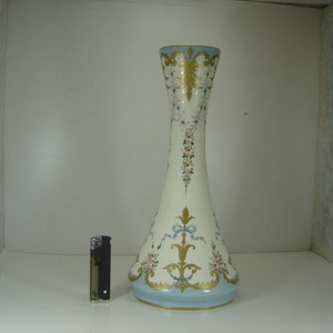 May include: A tall, white ceramic vase with a blue base and gold accents. The vase is decorated with floral and ribbon designs in shades of pink, blue, and gold.