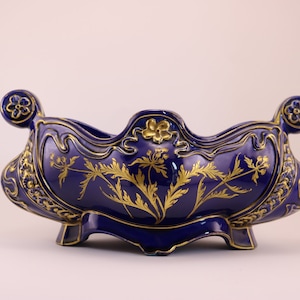 Puede incluir: Un cuenco de cer&aacute;mica azul y dorado con un dise&ntilde;o floral. El cuenco tiene un borde festoneado y dos asas. Est&aacute; decorado con pan de oro y tiene un patr&oacute;n floral.