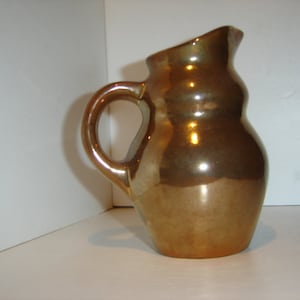 May include: A small, gold-colored ceramic pitcher with a rounded body and a slightly flared lip. The pitcher has a single handle and a smooth, glossy finish.