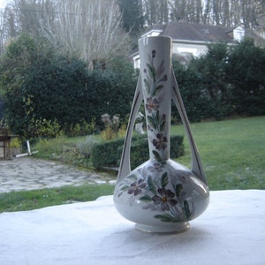 May include: A white ceramic vase with a floral design. The vase has two handles and a narrow neck. The flowers are painted in shades of pink, purple, and green.