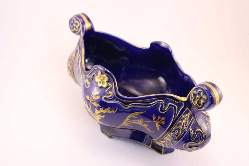 Puede incluir: Un cuenco de cer&aacute;mica azul cobalto con dise&ntilde;os florales y de volutas doradas. El cuenco tiene forma de barco y tiene dos asas.