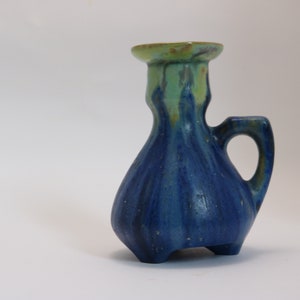 Puede incluir: Un pequeño jarrón de cerámica azul y verde con asa y tres patas. El jarrón tiene un esmalte que le da un aspecto moteado.
