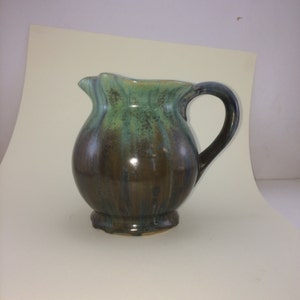May include: A small, green and brown ceramic pitcher with a rounded body and a handle. The pitcher has a glazed finish with streaks of green and brown.