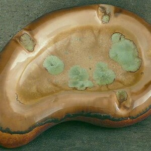 May include: A brown ceramic ashtray with a unique, organic shape and green and brown glaze. The ashtray has four indentations for cigarettes.