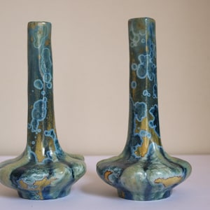 May include: A pair of small, blue and green ceramic vases with a gold glaze. The vases have a unique, organic shape and are perfect for displaying small flowers or greenery.