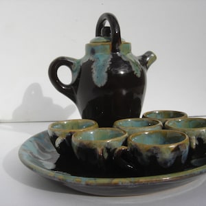 May include: A brown and green ceramic teapot with a matching tray and six cups. The teapot has a rounded body and a spout on the side. The cups are small and have a rounded shape. The tray is round and has a raised edge.