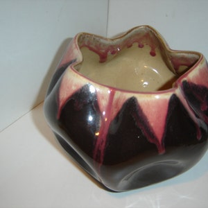 May include: A black ceramic planter with a unique, irregular shape. The planter has a beige interior and a pink glaze that drips down the sides.