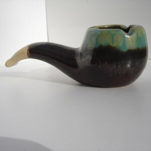 May include: A brown and green ceramic pipe ashtray. The pipe is glazed with a brown and green glaze. The bowl of the pipe is wide and round. The stem of the pipe is long and curved.