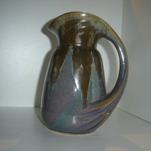 May include: A ceramic pitcher with a unique, curved handle. The pitcher is glazed in shades of brown, purple, and green.