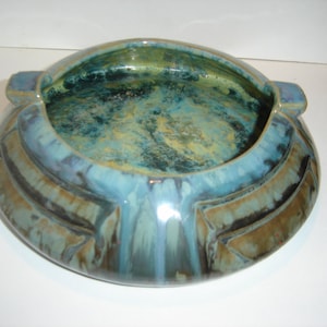 May include: A large, oval-shaped ceramic bowl with a blue, green, and brown glaze. The bowl has a wide, shallow opening and two small handles on the sides.