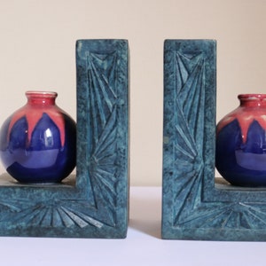 May include: A pair of blue and green ceramic bookends with a carved geometric design. Each bookend features a small, round vase with a blue and pink glaze.