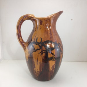 May include: A brown ceramic pitcher with a black design of stylized animals. The pitcher has a wide mouth and a handle.