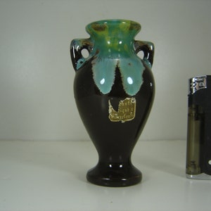 Puede incluir: Un pequeño jarrón de cerámica marrón oscuro con un esmalte verde y azul. El jarrón tiene dos asas y una etiqueta que dice "antiguo, Países Bajos".