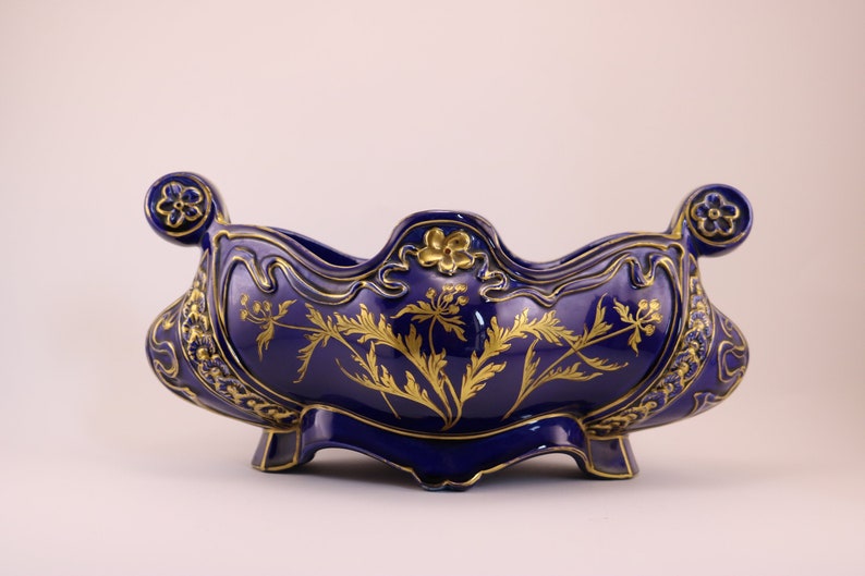 Puede incluir: Un cuenco de cer&aacute;mica azul y dorado con un dise&ntilde;o floral. El cuenco tiene un borde festoneado y dos asas. Est&aacute; decorado con pan de oro y tiene un patr&oacute;n floral.