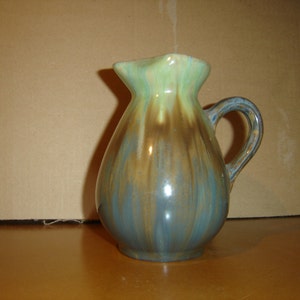 May include: A small, blue, green, and brown ceramic pitcher with a handle. The pitcher has a glazed finish and a rounded body.