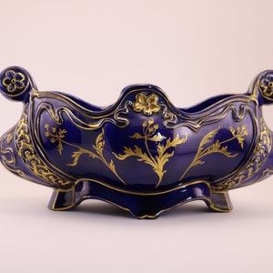 Puede incluir: Un cuenco de cer&aacute;mica azul cobalto con detalles florales dorados y adornos ornamentados. El cuenco tiene un borde festoneado y dos asas.