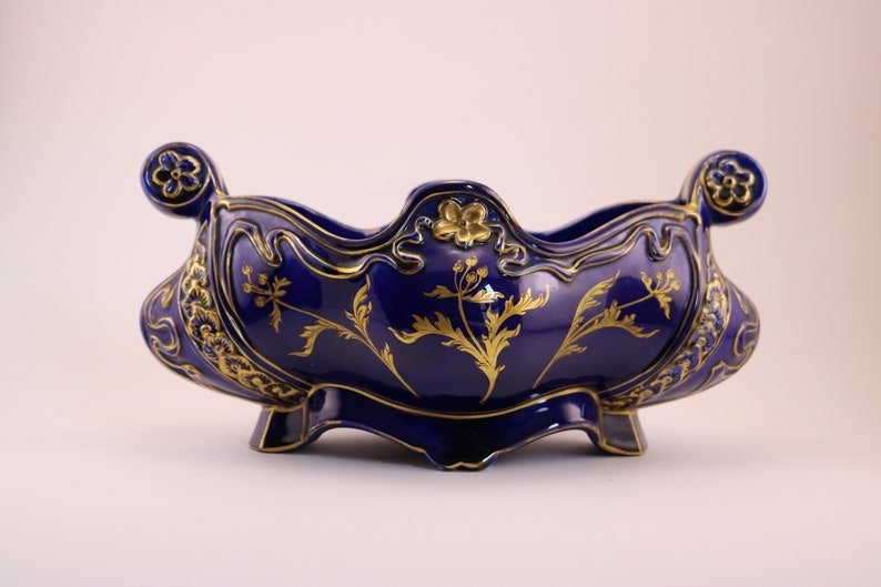 Puede incluir: Un cuenco de cer&aacute;mica azul cobalto con detalles florales dorados y adornos ornamentados. El cuenco tiene un borde festoneado y dos asas.