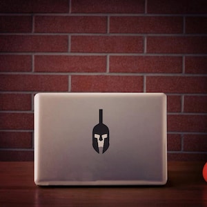 Può includere: Un computer portatile chiuso con un adesivo di un elmo spartano nero sul coperchio. Il computer portatile è appoggiato su una superficie di legno davanti a un muro di mattoni. Una palla rossa è appoggiata sulla superficie a destra del computer portatile.