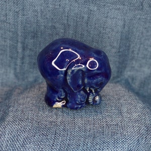 May include: A small, blue ceramic elephant figurine. The elephant is standing on all four legs and has its trunk curled up. The figurine is glazed in a deep blue color and has white accents.