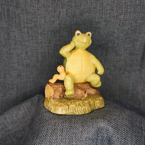 May include: A green ceramic turtle figurine sitting on a log with a yellow worm figurine. The turtle has a yellow shell and is sitting with its head tilted to the side. The figurine is on a base with green grass and brown dirt.